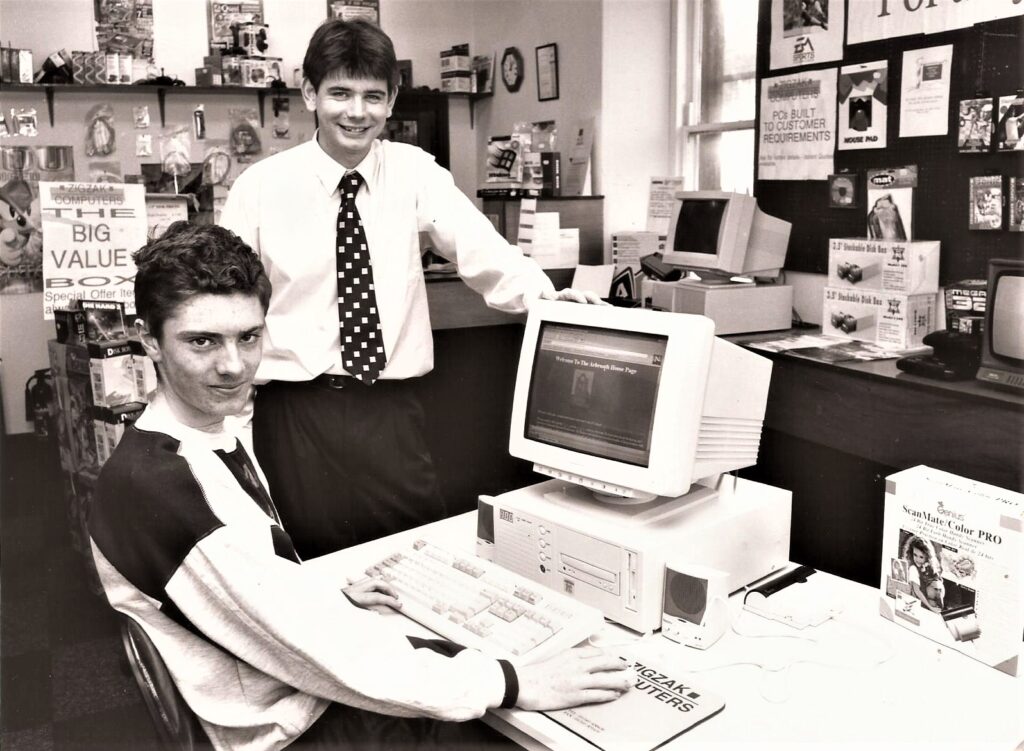 A person sitting at a 1990s style computer system while someone else stands next to them.