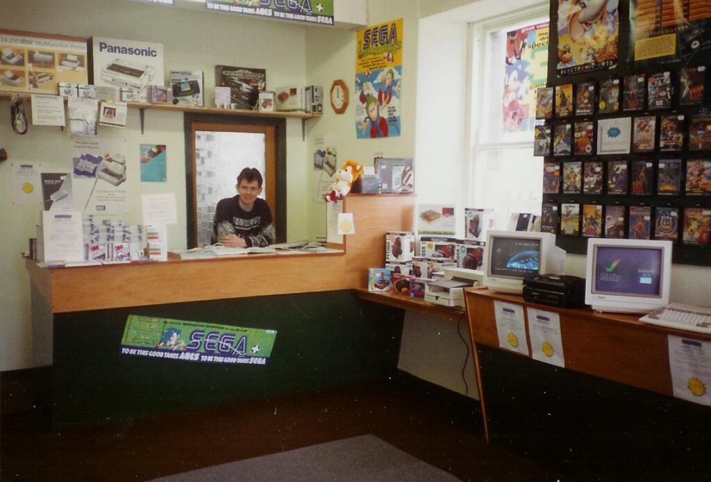 A picture of the inside of a computer shop from the mid 1990s, with a number of Sega games hanging on the wall and some demo equipment on a counter.