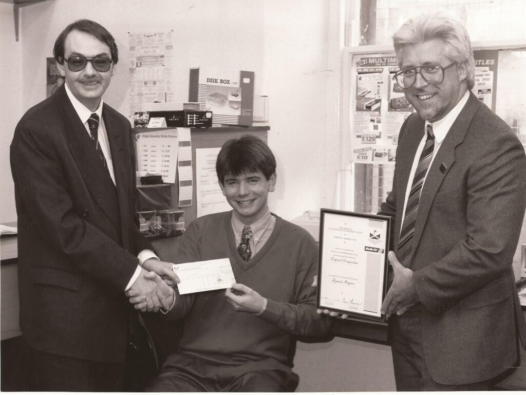 A cheque and certificate are presented to a seated man by two others standing either side.